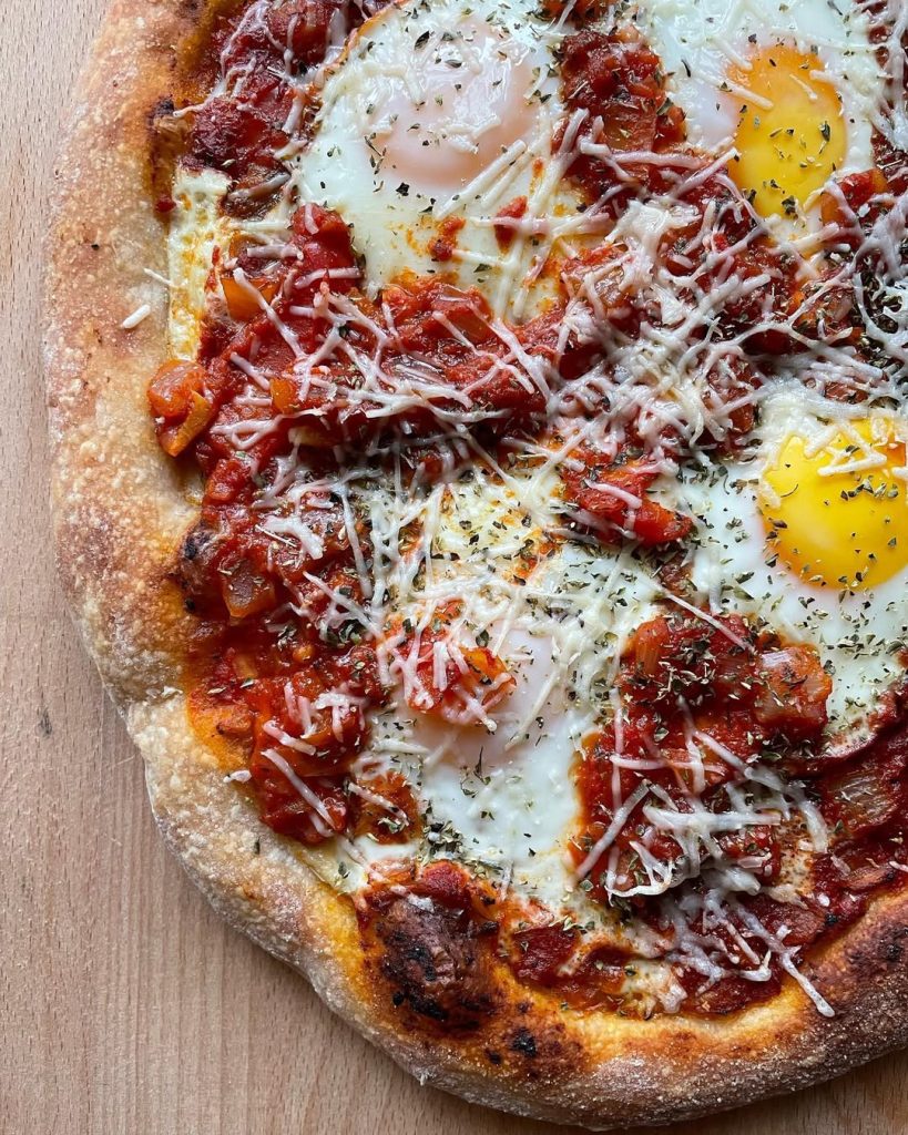 Shakshuka-inspired pizza, featuring baked eggs nestled in a rich spiced tomato and capsicum sauce, sprinkled with grated Grana Padano and dried herbs.