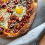 Shakshuka-inspired pizza, featuring baked eggs nestled in a rich spiced tomato and capsicum sauce, sprinkled with grated Grana Padano and dried herbs.