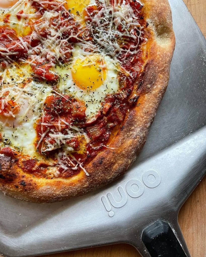 Shakshuka-inspired pizza, featuring baked eggs nestled in a rich spiced tomato and capsicum sauce, sprinkled with grated Grana Padano and dried herbs.