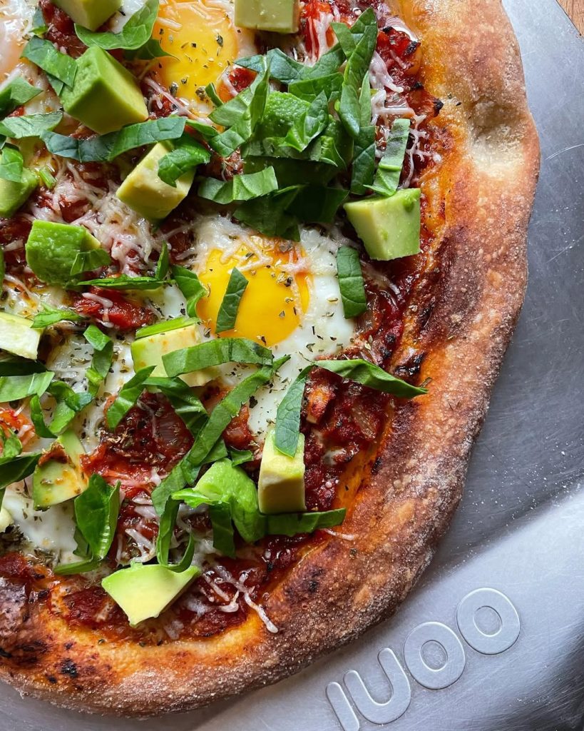 Shakshuka-inspired pizza, featuring baked eggs nestled in a rich spiced tomato and capsicum sauce, sprinkled with grated Grana Padano and dried herbs. Topped with avocado and spinach.