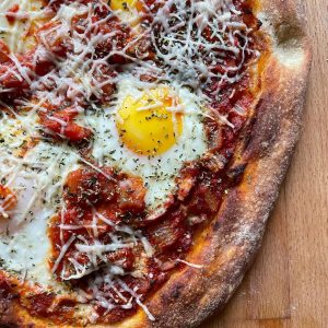 Shakshuka-inspired pizza, featuring baked eggs nestled in a rich spiced tomato and capsicum sauce, sprinkled with grated Grana Padano and dried herbs.