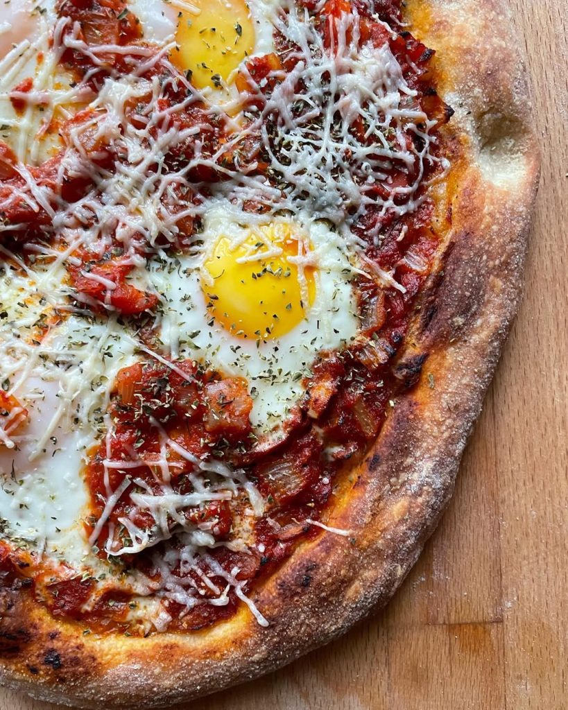 Shakshuka-inspired pizza, featuring baked eggs nestled in a rich spiced tomato and capsicum sauce, sprinkled with grated Grana Padano and dried herbs.