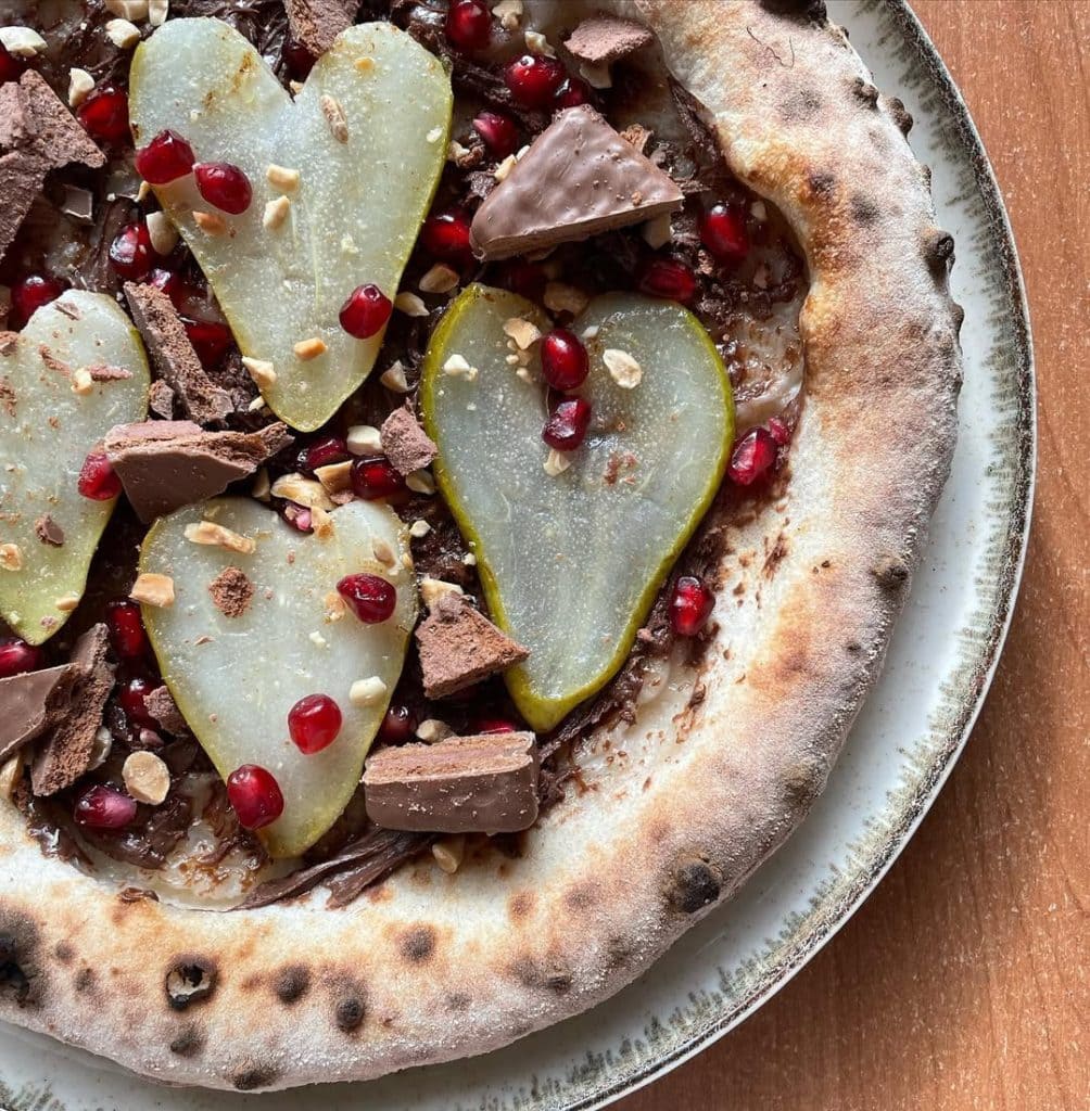 Pizza with Nutella, pear slices, crushed Tim Tam biscuits, chopped peanuts, and bright pomegranate arils on a golden crust, served warm on a wooden board.