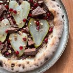 Pizza with Nutella, pear slices, crushed Tim Tam biscuits, chopped peanuts, and bright pomegranate arils on a golden crust, served warm on a wooden board.
