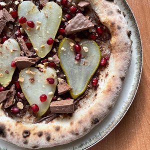 Pizza with Nutella, pear slices, crushed Tim Tam biscuits, chopped peanuts, and bright pomegranate arils on a golden crust, served warm on a wooden board.
