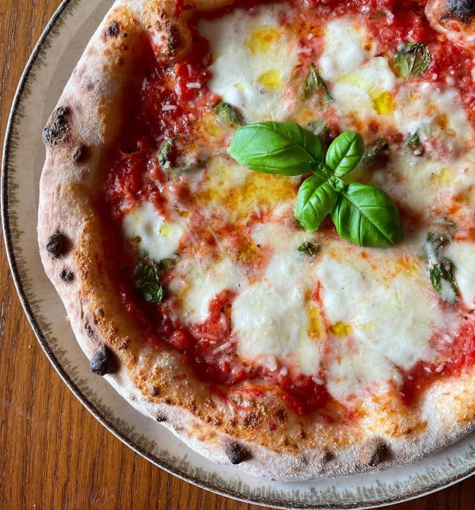 Neapolitan-style Margherita pizza topped with tomato sauce, melted mozzarella, basil leaves added before and after baking, and a drizzle of olive oil, showcasing a puffy, golden crust from a long room-temperature fermentation