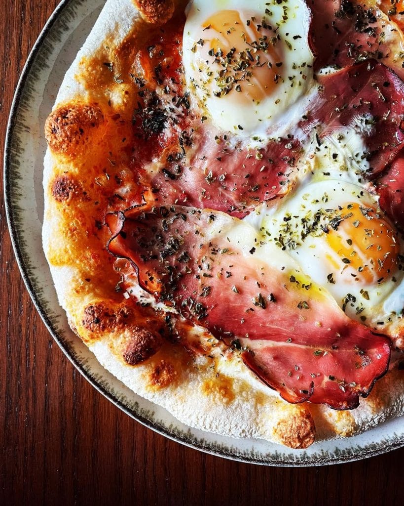 Aussie-style pizza topped with sunny-side-up eggs, crispy-edged prosciutto, melted mozzarella, and a sprinkle of oregano, with a drizzle of olive oil.