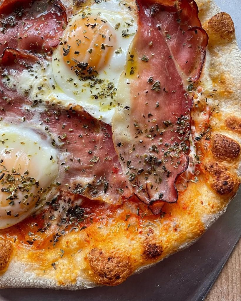 Aussie-style pizza topped with sunny-side-up eggs, crispy-edged prosciutto, melted mozzarella, and a sprinkle of oregano, with a drizzle of olive oil.