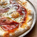 Aussie-style pizza topped with sunny-side-up eggs, crispy-edged prosciutto, melted mozzarella, and a sprinkle of oregano, with a drizzle of olive oil.