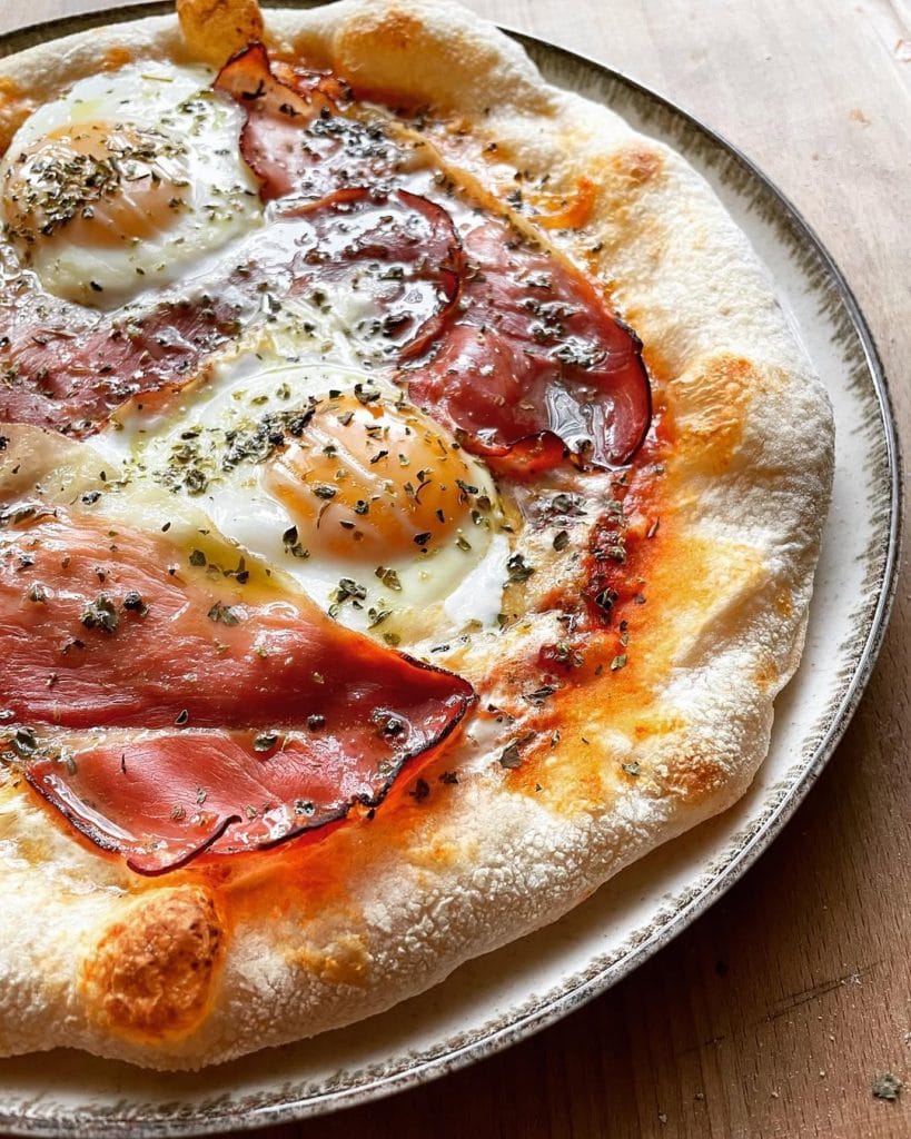 Aussie-style pizza topped with sunny-side-up eggs, crispy-edged prosciutto, melted mozzarella, and a sprinkle of oregano, with a drizzle of olive oil.