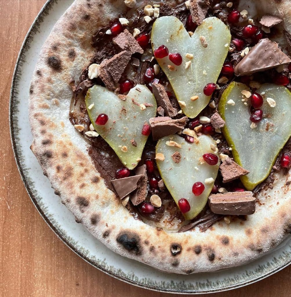 Pizza with Nutella, pear slices, crushed Tim Tam biscuits, chopped peanuts, and bright pomegranate arils on a golden crust, served warm on a wooden board.