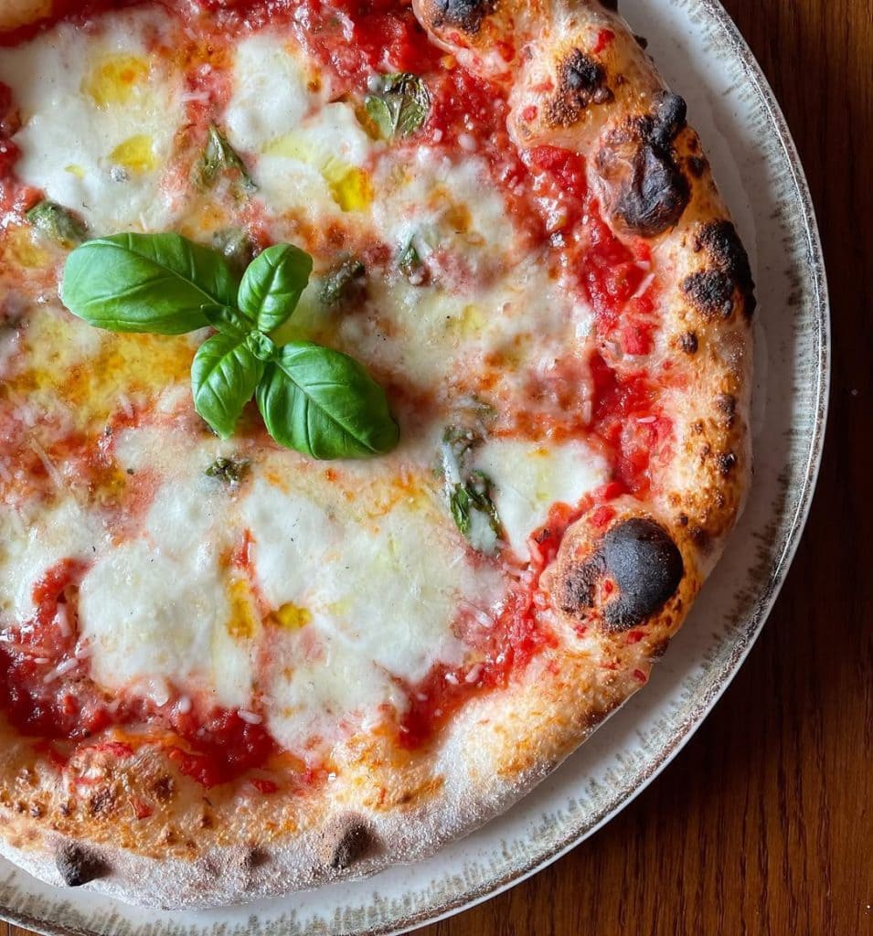 Neapolitan-style Margherita pizza topped with tomato sauce, melted mozzarella, basil leaves added before and after baking, and a drizzle of olive oil, showcasing a puffy, golden crust from a long room-temperature fermentation