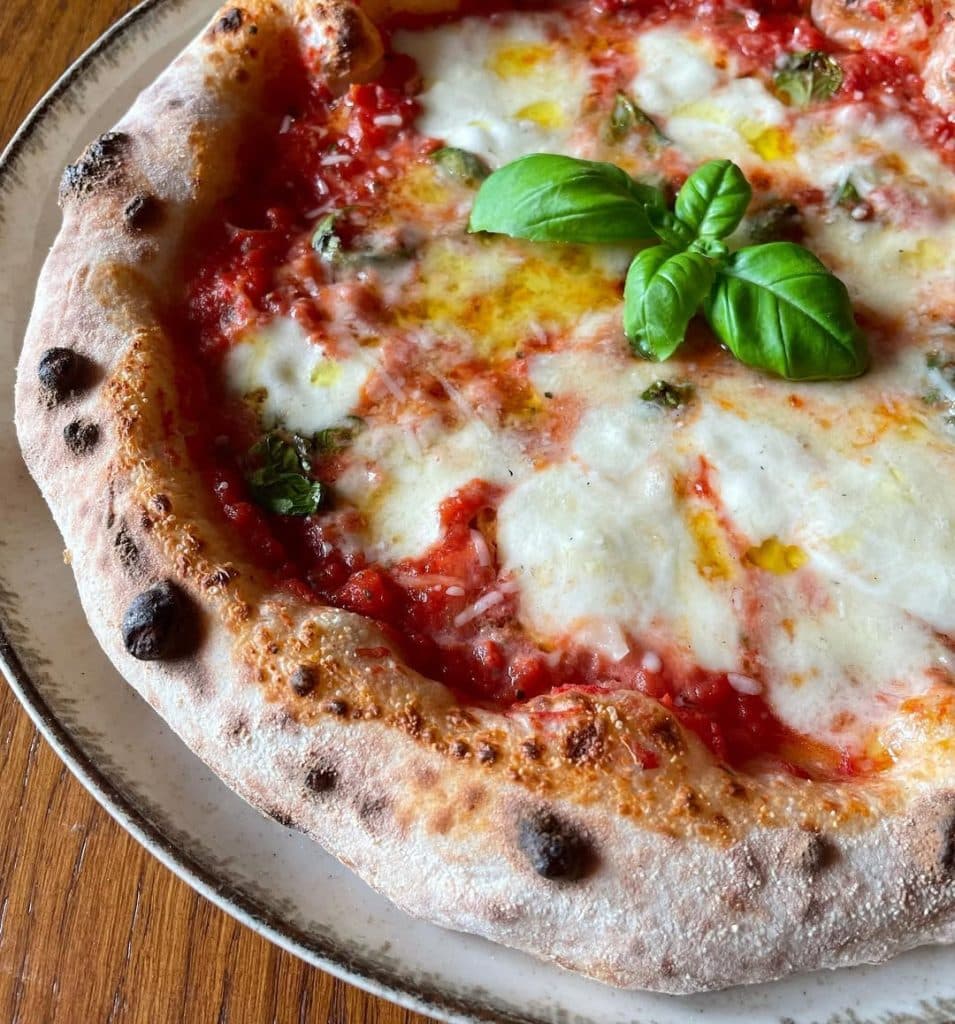 Neapolitan-style Margherita pizza topped with tomato sauce, melted mozzarella, basil leaves added before and after baking, and a drizzle of olive oil, showcasing a puffy, golden crust from a long room-temperature fermentation
