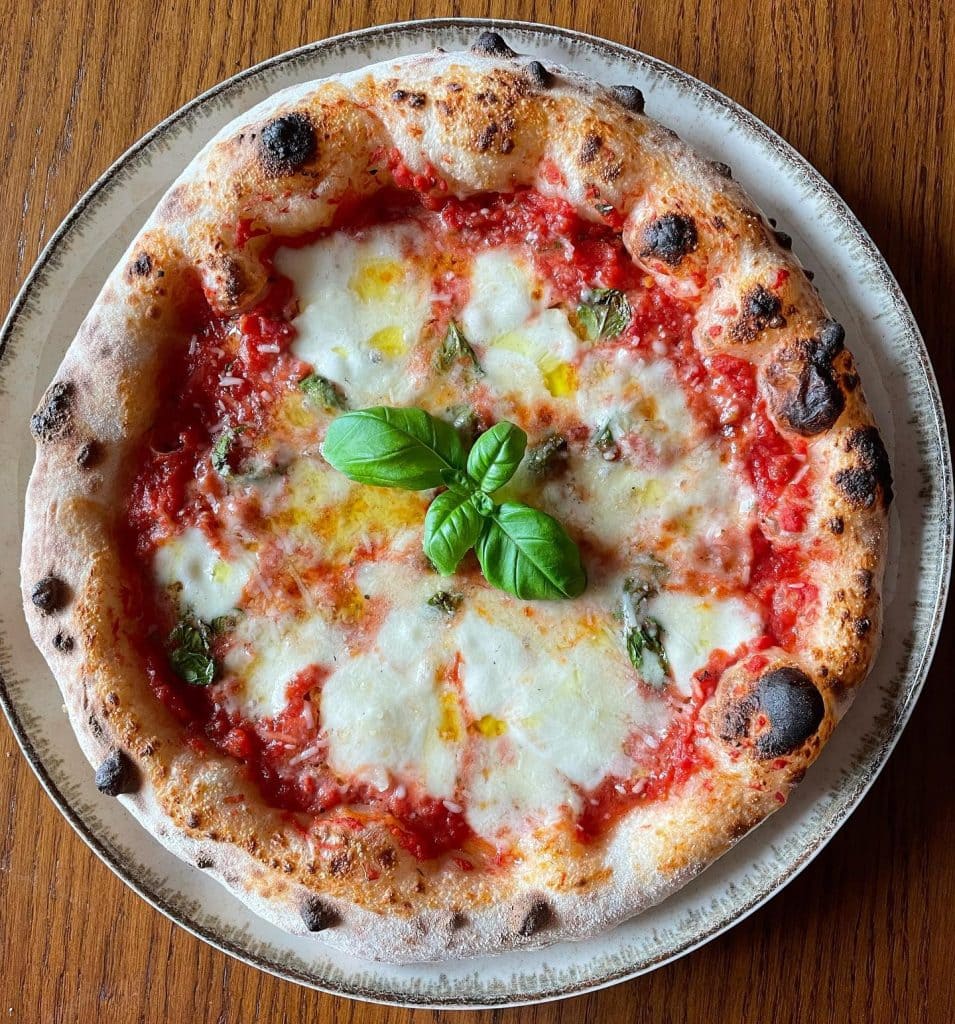 Neapolitan-style Margherita pizza topped with tomato sauce, melted mozzarella, basil leaves added before and after baking, and a drizzle of olive oil, showcasing a puffy, golden crust from a long room-temperature fermentation